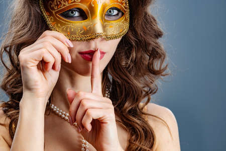 Brunette woman in a carnival mask on a blue background touching her finger to her lips looking at the camera closeupの写真素材