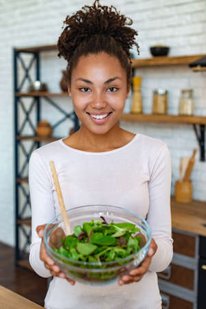 Pretty mulatto woman at home showing healthy food, holds in her hands a salad - Imageの写真素材