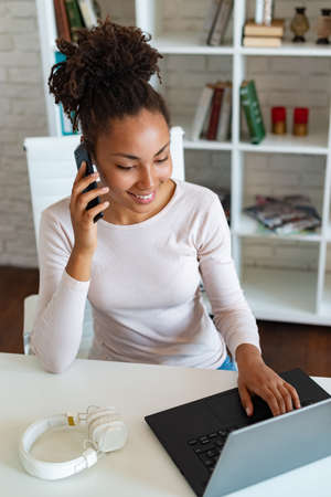 Smiling mulatto woman working on laptop in the office and talking on a smartphone . - Imageの写真素材