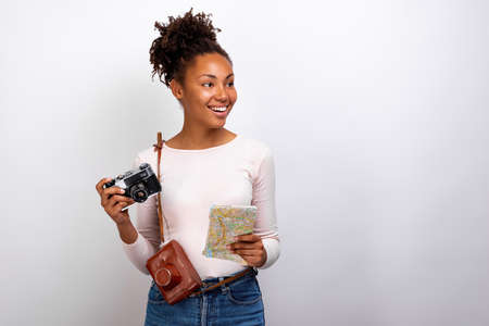 Studio portrait of a happy mulatto traveller girl with photo camera and a map in her hands and looking sideways- Imageの写真素材