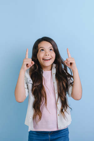 Excited girl standing over blue studio background and pointing up her fingers to copyspace looking upの写真素材