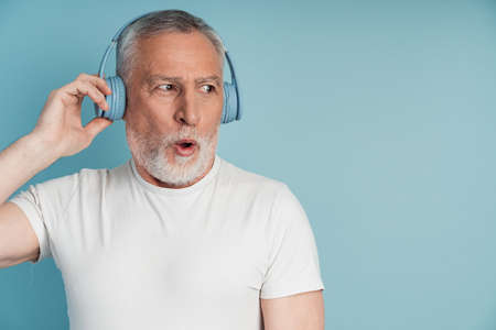 Surprised man in headphones posing on blue wall background, isolated. Positive senior man listening to music on headphonesの写真素材