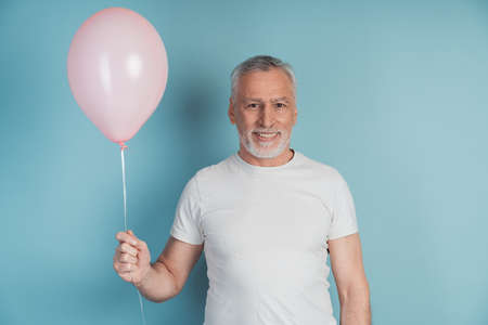 Happy retired man in white t-shirt holding pink balloon while posing in studio. Senior, smiling man over blue background, copy space, place for textの写真素材