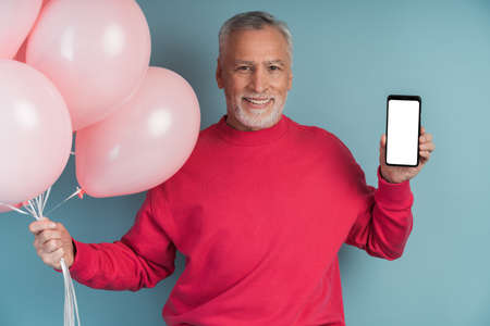 Man with balloons wearing bright pullover posing on blue background. Nice man holding a phone, showing a blank, black phone screen, copy space, place for textの写真素材