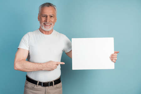 Smiling, pleasant, senior man holds a white piece of paper, points to it. Isolated on blue background man holding blank white piece of paper, copy space, place for advertising.の写真素材
