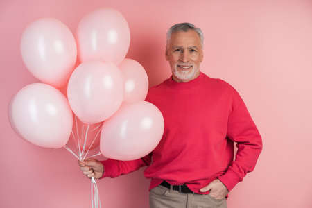 Happy man celebrating holding pink balloons. Cheerful, senior man in a bright pullover over pink backgroundの写真素材