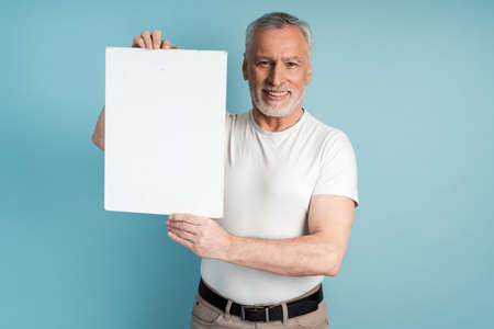 Smiling retiree with a beard holding a blank sheet of paper on a blue background. Man isolated on copy space.の写真素材