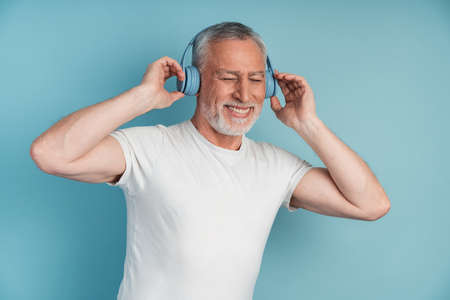 Mature gray-haired man has a great time, brings in headphones and listens to music. Positive gentleman enjoys music and dancing on a blue backgroundの写真素材