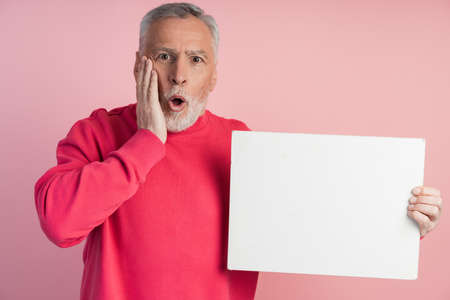 Surprised senior man holding a white sheet of paper. Isolated on pink background man holding blank white sheet of paper, copy space, place for advertising.の写真素材