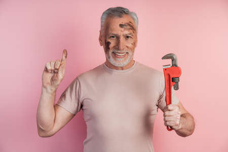 Positive, senior man with gray hair and a beard holds a tool in his hand and raised his index finger up. Man isolated on pink background.の写真素材