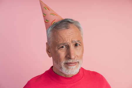 Sad, upset senior man in a festive hat on a pink background. Dissatisfied man isolated on copy space.の写真素材