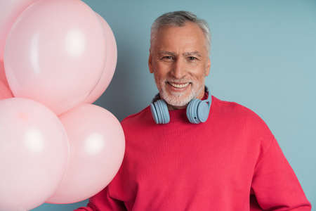 Handsome, smiling man with gray hair wears headphones, holding balloons. Attractive senior man on a blue background.の写真素材