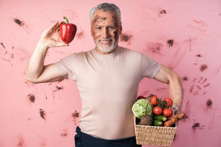 Smiling, elderly man holds a basket of vegetables and holds a red pepper in his other hand. Farmer on a dirty pink background.の写真素材