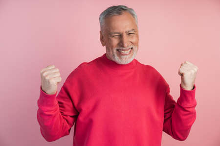 Joyful, senior man posing on a pink background, he loves the gesture of victory. The man is very happy, he closed his eyes and rejoices in the victory, clenched his fists.の写真素材