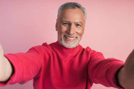 Close-up shot of a senior man's hands holding his phone, taking a selfie, on a pink background. Smiling man isolated on pink background.の写真素材