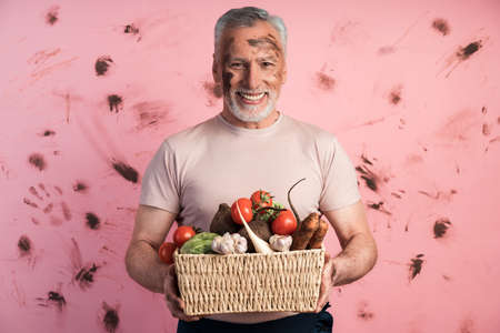 Smiling, positive senior man holding a basket of fresh vegetables against a dirty pink wall. The concept of farming, household.の写真素材