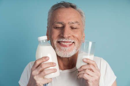 Cute grandfather with gray hair and beard holds a bottle of milk and a glass, wears an apron. Nice man on a blue background closed his eyes, touches the bottle and a glass of his face, copy the space, place for text.の写真素材