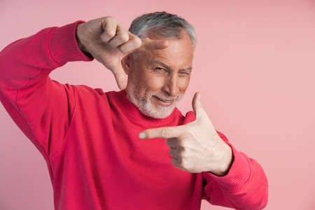Gray-bearded man makes a frame with his fingers, photo. Attractive, senior man posing on a pink background.の写真素材