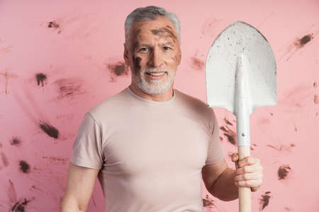 Positive man with a shovel on a background of a dirty pink wall. Senior man, farmer on copy space.の写真素材