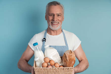 Handsome, in years, a senior man holding a basket with food bread, milk, eggs. Smiling man on a blue background.の写真素材