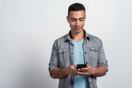 Half-length portrait of a mulatto man standing against the white background holding mobile phone in his handsの写真素材