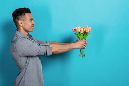 Happy mulatto man holding his hands straigth himself with  a tulips bouquet  against a blue studio background .- Imageの写真素材