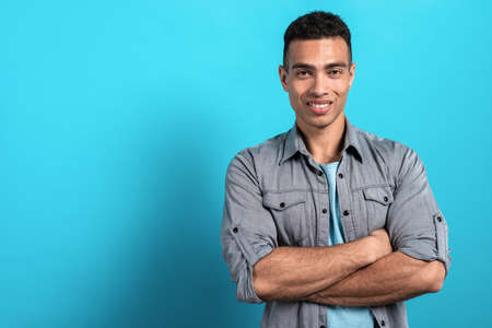 Young happy mulatto man folded his hands and standing against a blue stidio background. - Imageの写真素材
