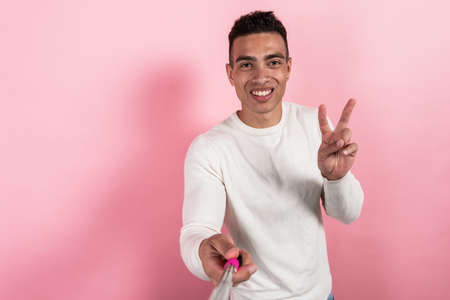 Happy mulatto man taking a self portrait with selfie stick ahnd showing a victoty gesture against a pink background. - Imageの写真素材
