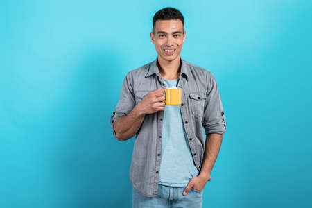 Pretty man smiling holding a yellow cup in his hand standing against a blue background- Imageの写真素材