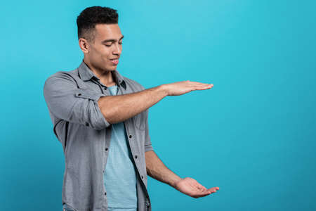 Smiling mulatto man shows big size with both hands on blue studio background demonstrate large space or holding object, copy spaceの写真素材