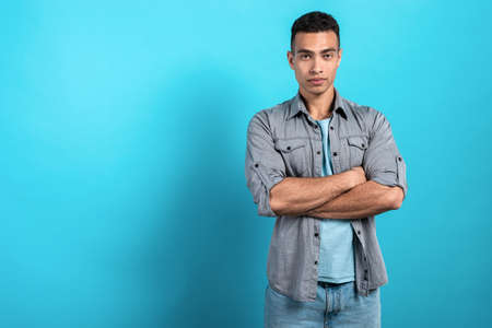 Young serios mulatto man folded his hands and standing against a blue stidio background. - Imageの写真素材
