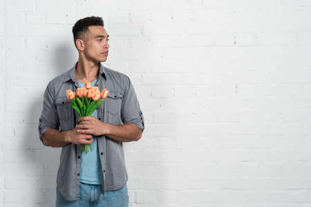 Handsome mulatto man standing against a white brick wall holding a tulip bouquet in his hands and looking away. - Copy spaceの写真素材