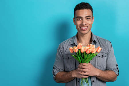 Portrait of happy mulatto man standing against a blue wall holding a tulip bouquet in his hands .- Imageの写真素材