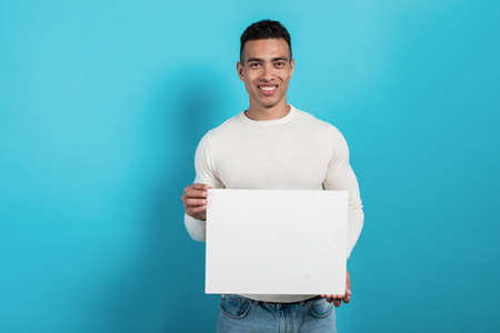 Happy young  mulatto man holds in his hands a white blank sheet. Advertising space.- Imageの写真素材