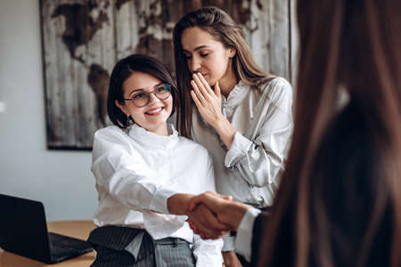 Smiling girl in glasses shakes hands with her colleague while her assistant says something in her...