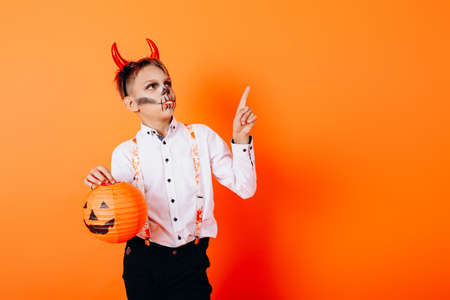 Boy in Devil masquerade makeup holding a pumpkin and pointing up. Halloween conceptの写真素材