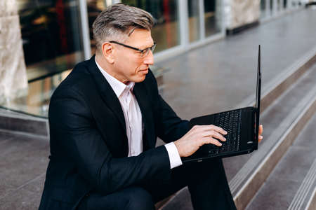 Senior businessman sitting on pavement, holding a laptop and typing. - Imageの写真素材