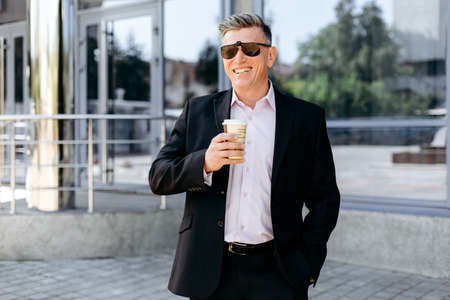 Portrait of  senior businessman standing on pavement holding a cup of coffee and smiling . - Imageの写真素材