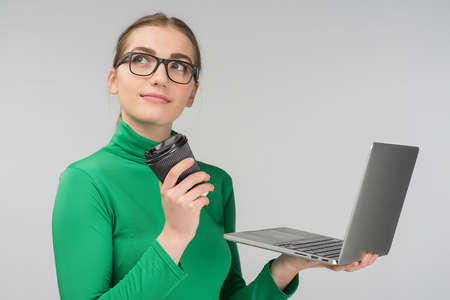 Dreamy woman  holds a cup of coffee and laptop in her hands  while standing against on the white background.の写真素材