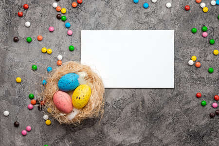 On a gray plaster background lies a bird's nest with colorful eggs, a blank, easter card and sweetsの写真素材