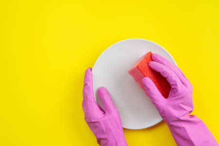Closeup hands wearing in latex gloves holding a kitchen sponge and plate.  Top view on yellow  background washing dishesの写真素材