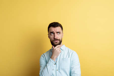 Studio male face portrait. Man toushes himself chin and frown seriously looking up. Yellow background. Copyspaceの写真素材