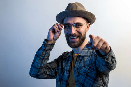 Portrait of bearded man in hat and touches his glasses fun smiling and pointing at the cameraの写真素材