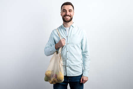 A young guy in a blue shirt stands and holds 
 reusable mesh bag with fruits. The concept of zero waste.の写真素材