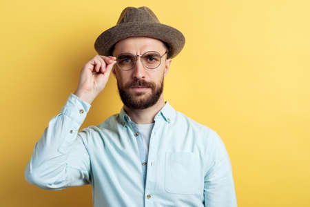 Portrait of brunette man with beard wearing in shirt, hat and glasses. He frown eyesbrows and looking at the camera touching glassesの写真素材