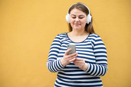 Pretty woman with a thin weight in headphones in a striped jacket on a background of orange wallの写真素材