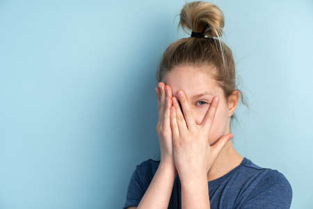 Afraid teenage girl who looks through her fingers. Teen girl covering face with hands isolated on blue background, copy space.の写真素材