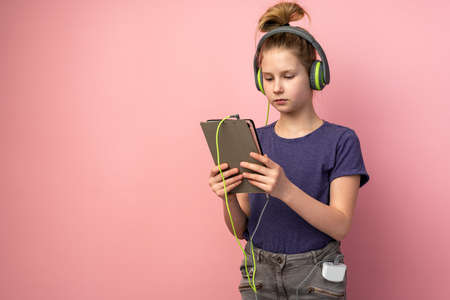 Serious teenage girl in headphones and tablet in hands on a pink background. A cute child is learning or viewing something on the device.の写真素材