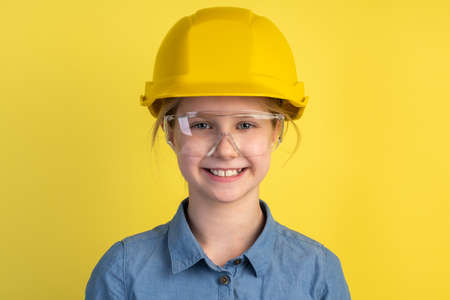 Cheerful, smiling child in a helmet and construction glasses on a yellow background. Girl isolated...