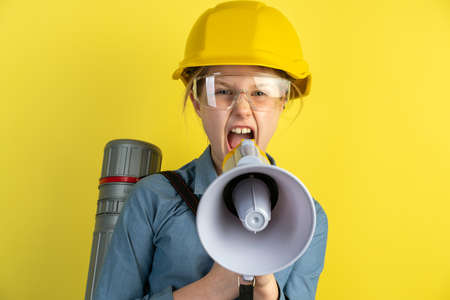 Portrait of a teenage girl in a helmet, a loudspeaker in her hands. Girl isolated on a yellow background, shouting through a megaphone.の写真素材